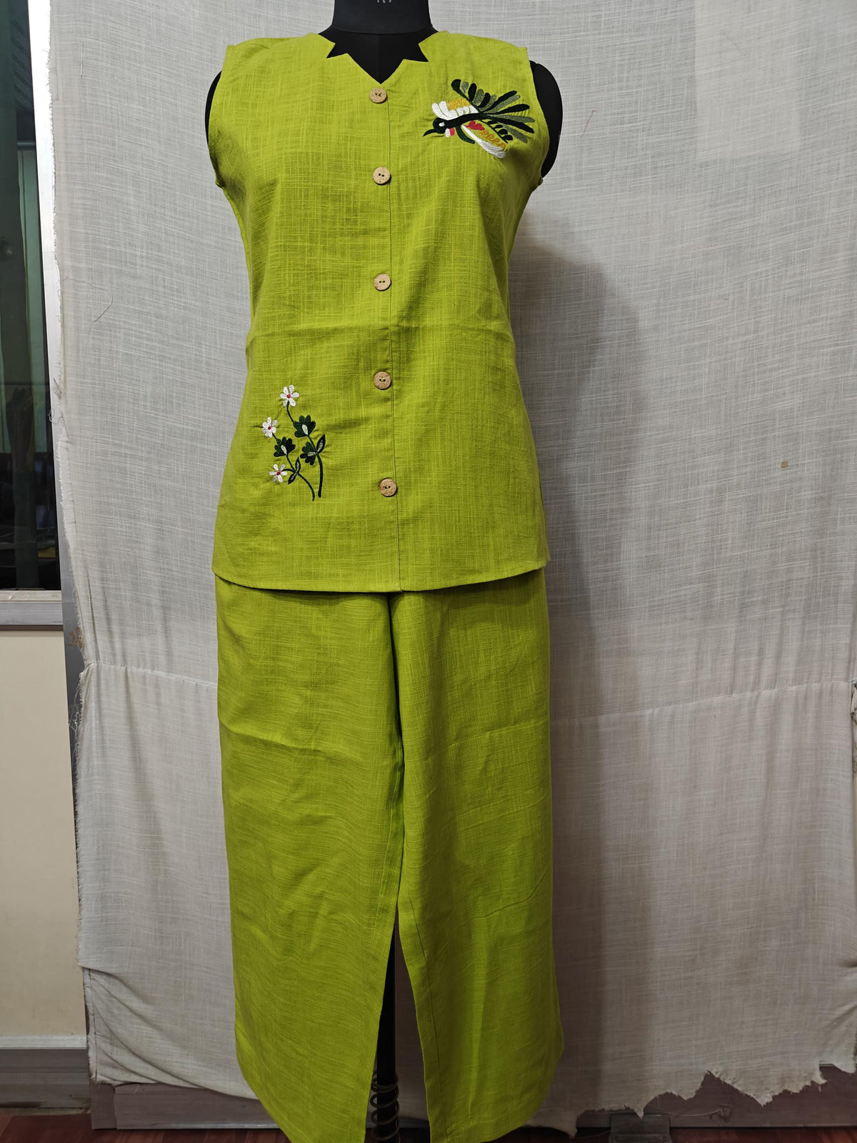 Puede incluir: Un conjunto de dos piezas verde lima en un maniquí. La parte superior sin mangas tiene una botonadura y detalles bordados de pájaros y flores. Los pantalones a juego son de pernera ancha. El conjunto está hecho de tela texturizada.