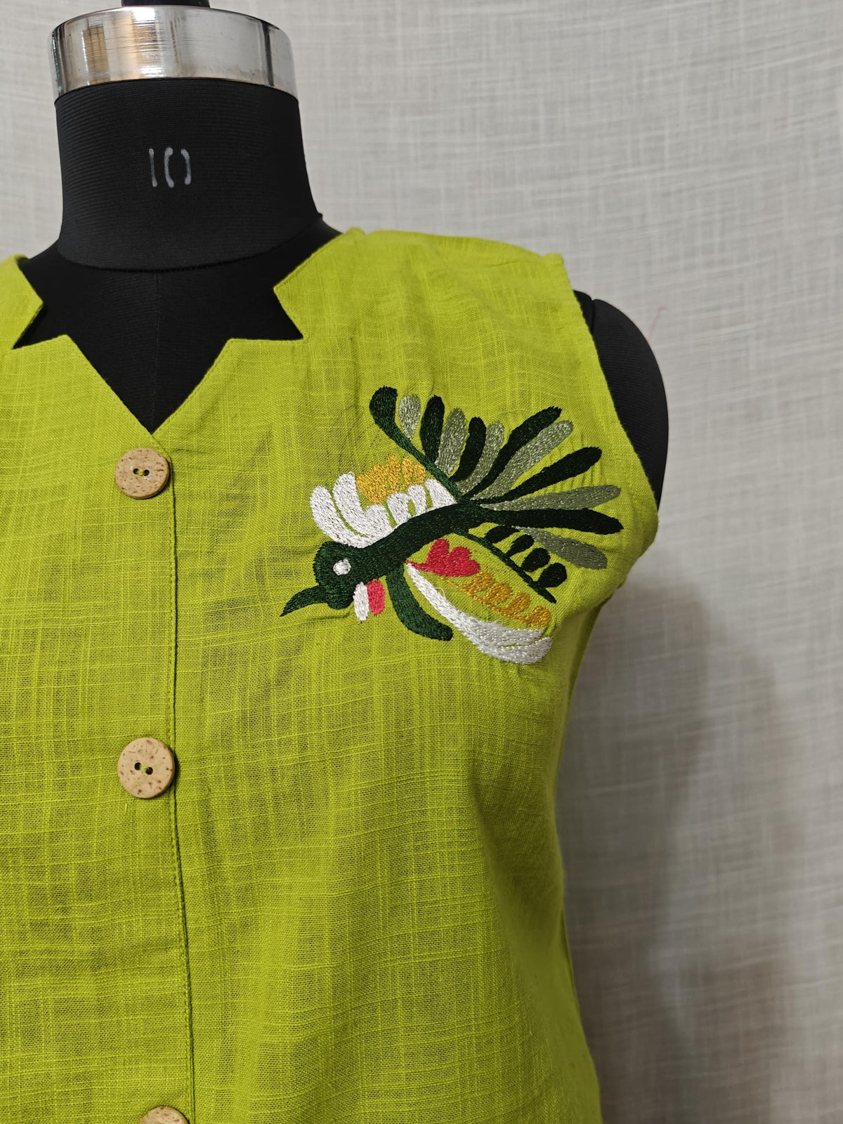 Puede incluir: Un chaleco sin mangas de color verde lima con cuello negro y botones de madera. El chaleco presenta un diseño de pájaro bordado en blanco, verde, amarillo y rojo. La tela tiene una apariencia texturizada.