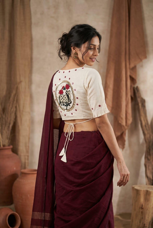 May include: A cream-coloured blouse featuring a central circular patch with a black cow illustration and red floral accents, surrounded by gold sequins. The blouse has short sleeves with pleated cuffs and a tie-back closure, paired with a dark maroon sari.