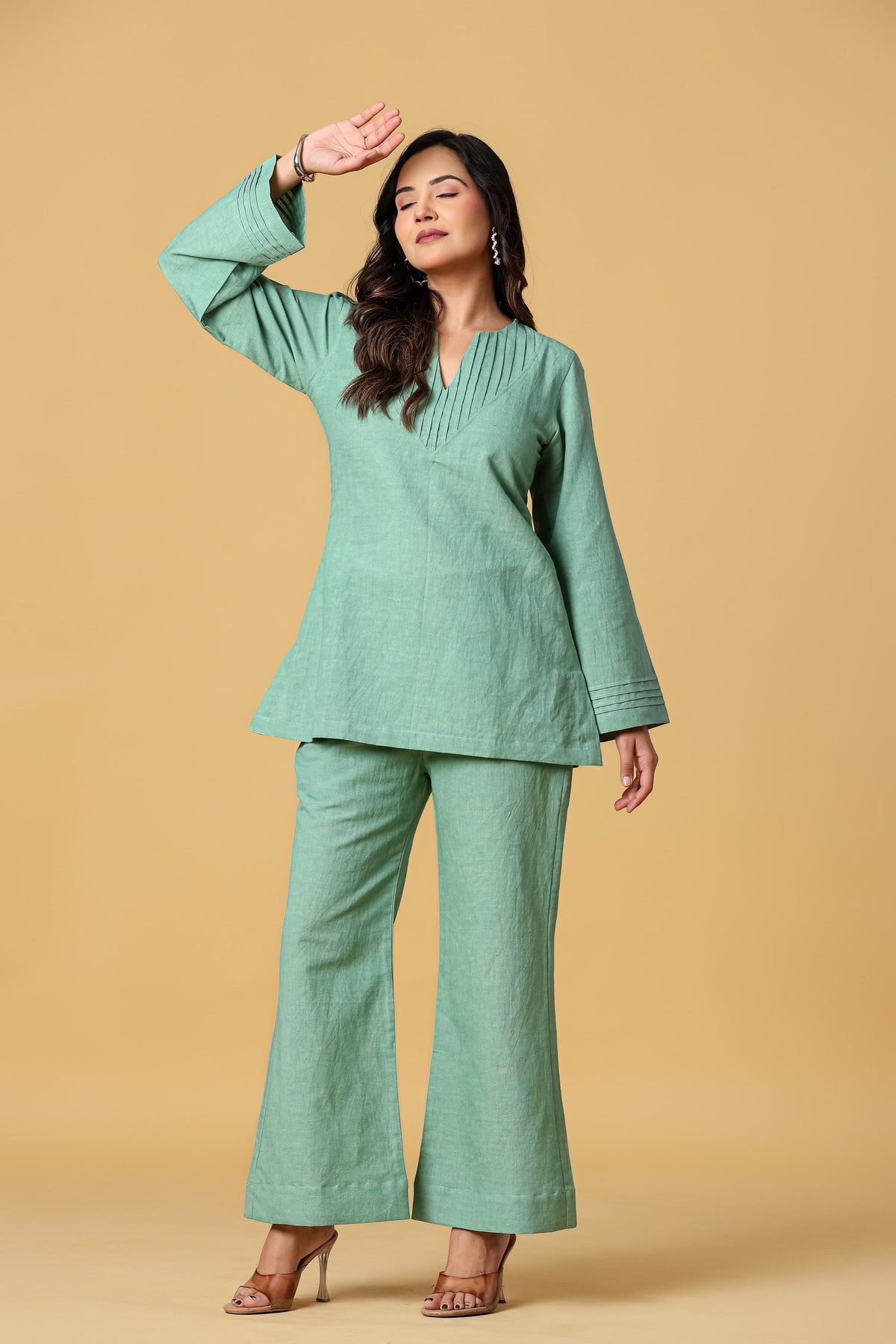 May include: A sage green two-piece set consisting of a long-sleeved tunic and wide-leg trousers. The tunic features a V-neckline with vertical pleat detailing and flared sleeves with horizontal stitched cuffs. The fabric has a textured finish.