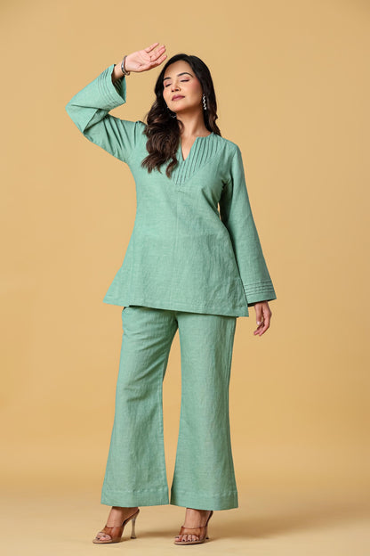 May include: A sage green two-piece set consisting of a long-sleeved tunic and wide-leg trousers. The tunic features a V-neckline with vertical pleat detailing and flared sleeves with horizontal stitched cuffs. The fabric has a textured finish.