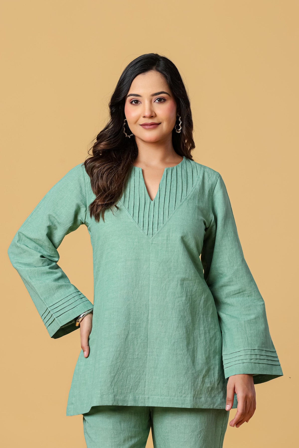 May include: A sage green long-sleeved tunic featuring a V-neckline with vertical pleat detailing. The garment includes matching pleats at the cuffs of the sleeves and a straight hemline, crafted from a solid-colored fabric with a visible texture.
