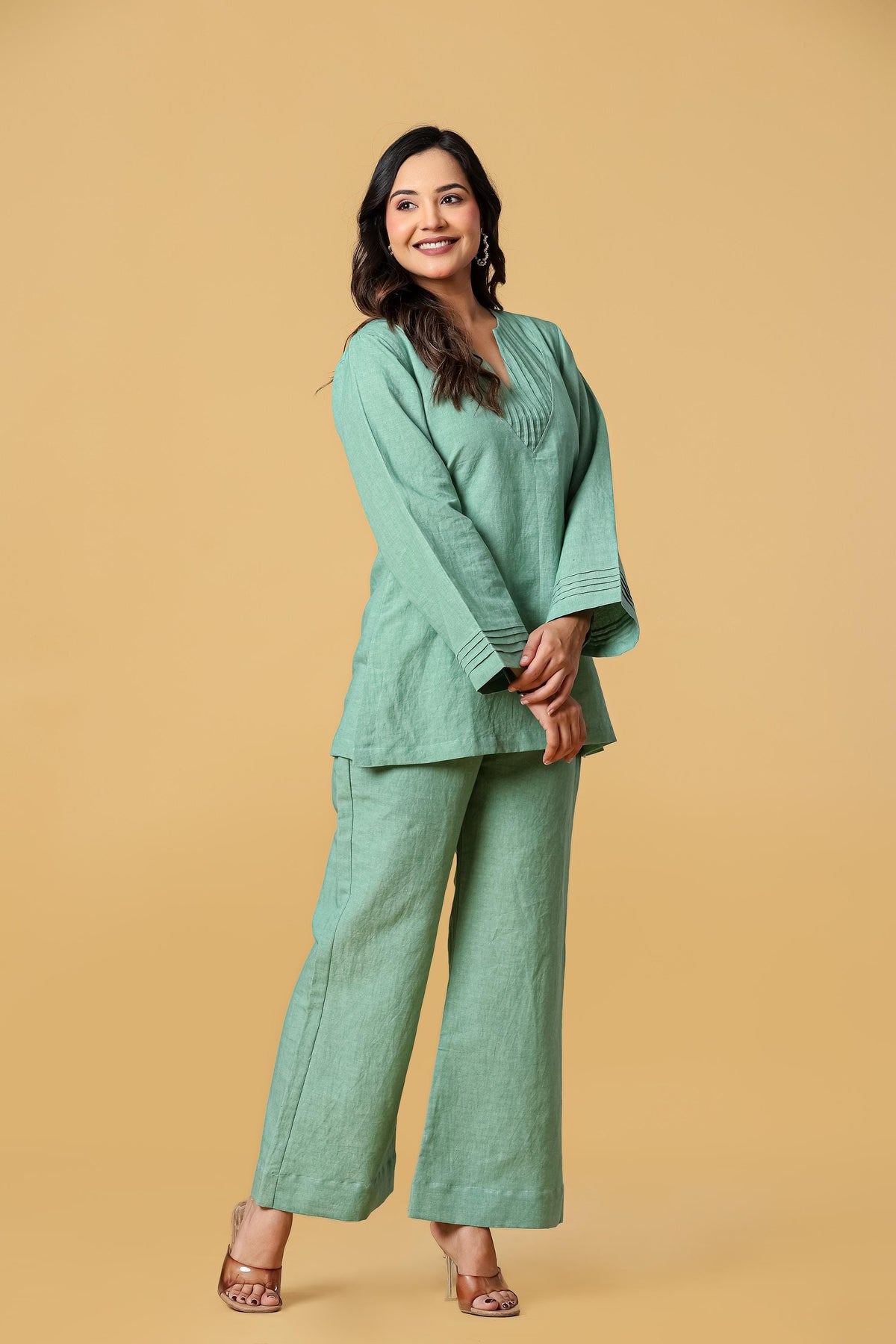 May include: A sage green two-piece set consisting of a long-sleeved tunic and matching wide-leg trousers. The tunic features a split V-neckline with vertical pleat detailing and matching pleats at the flared sleeve cuffs, creating a cohesive look.