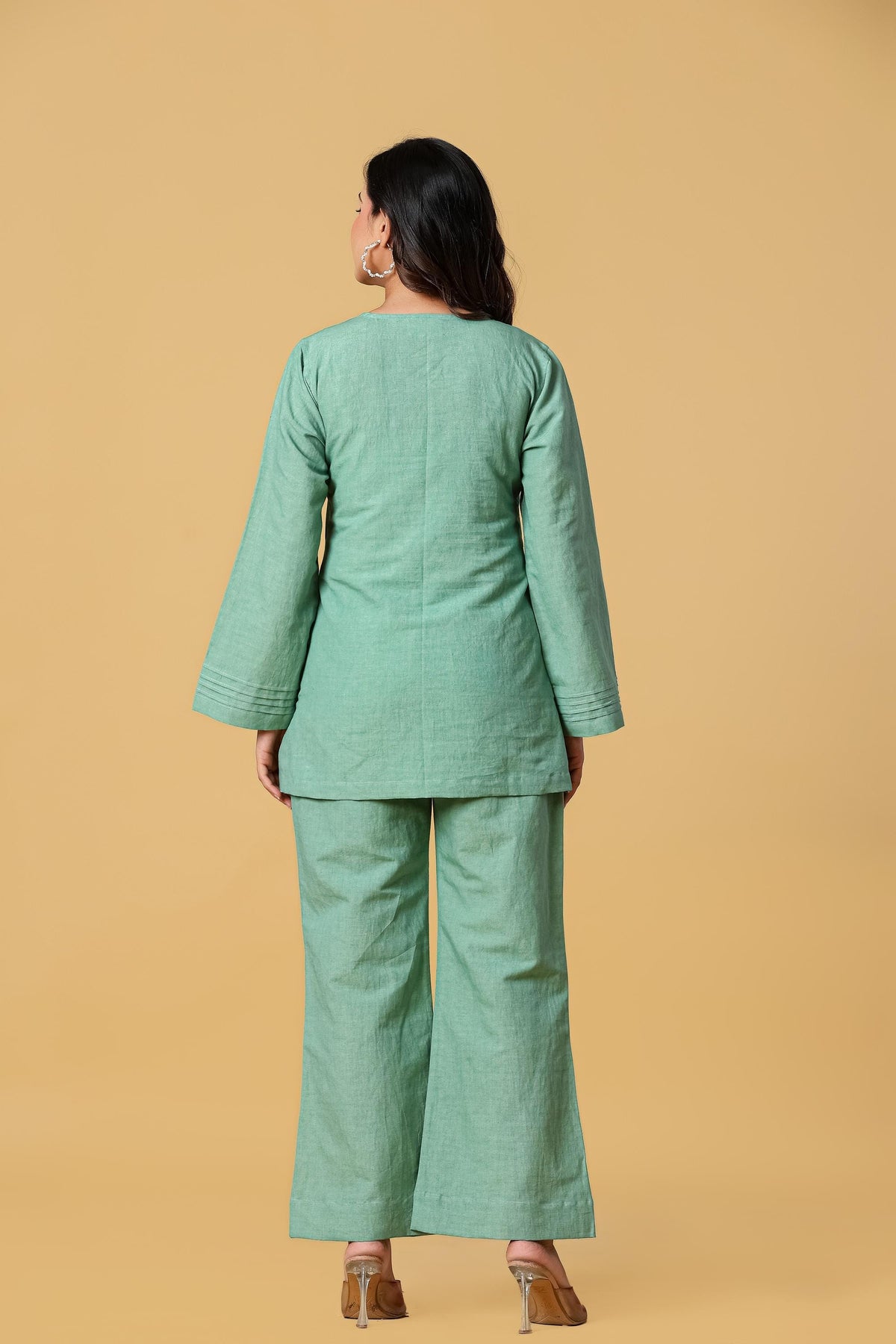 May include: A sage green two-piece set consisting of a long-sleeved tunic and matching wide-leg trousers. The tunic features a round neckline and subtle pleated detailing at the sleeve cuffs. The fabric has a visible textured weave throughout.
