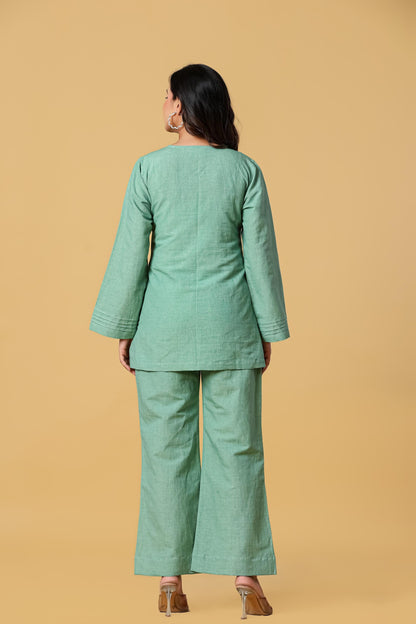 May include: A sage green two-piece set consisting of a long-sleeved tunic and matching wide-leg trousers. The tunic features a round neckline and subtle pleated detailing at the sleeve cuffs. The fabric has a visible textured weave throughout.