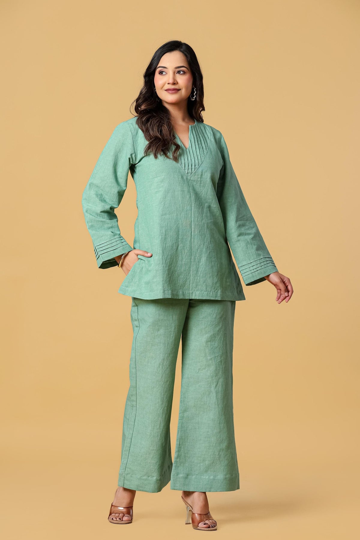 May include: A sage green two-piece set consisting of a long-sleeved tunic and wide-leg trousers. The tunic features a V-neckline with vertical pleat detailing and matching pleats on the sleeve cuffs. The trousers have a straight-leg silhouette.