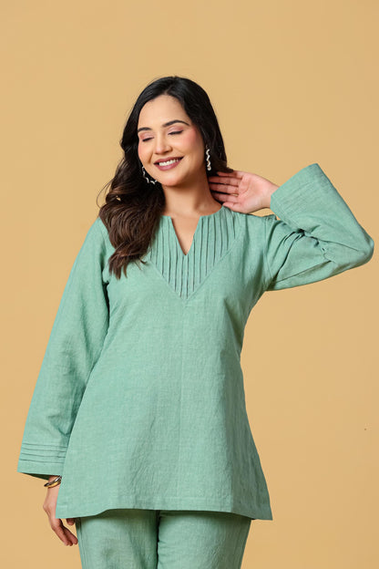 May include: A sage green long-sleeved tunic featuring a V-neckline with vertical pleat detailing at the chest. The garment has long sleeves with triple-stitched cuffs and a straight hemline, crafted from a textured fabric with a solid color finish.