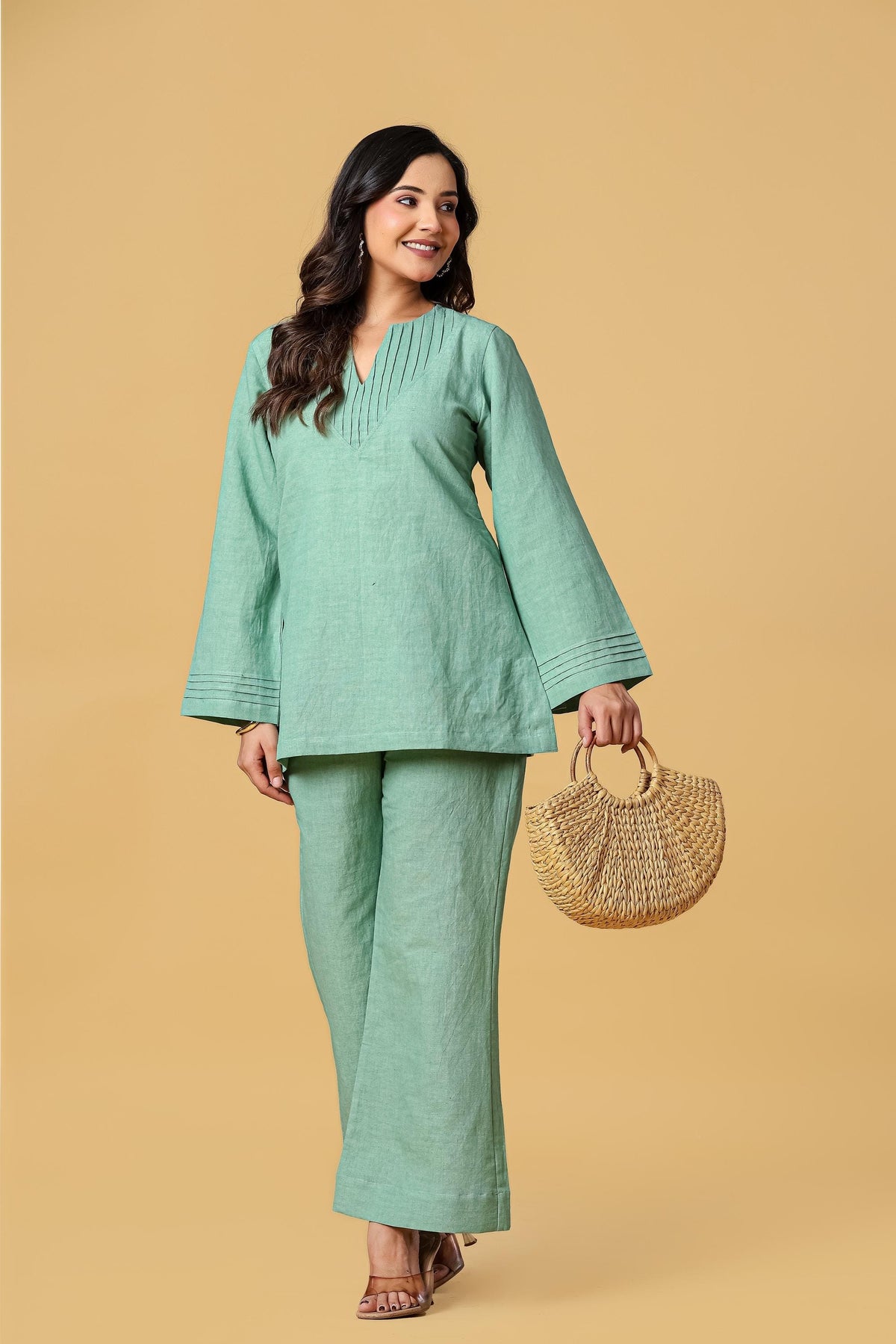 May include: A sage green long-sleeved tunic with a V-neckline featuring vertical pleats and matching wide-leg trousers. The tunic has pleated cuffs on the sleeves. A woven straw handbag with round handles is held in one hand.