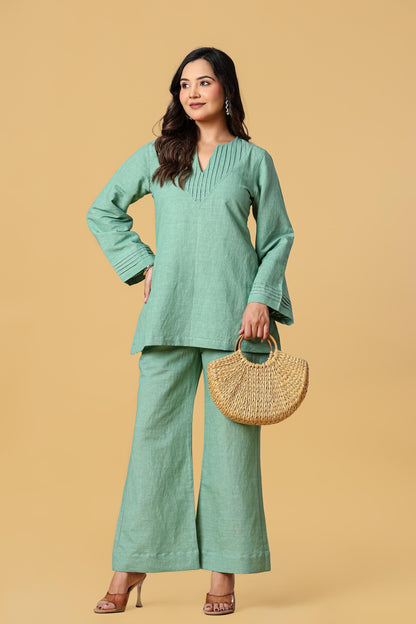 May include: A sage green long-sleeved tunic with a V-neckline featuring vertical pleats and matching wide-leg trousers. The outfit is paired with a semi-circular woven straw handbag with two round handles, creating a coordinated ensemble.