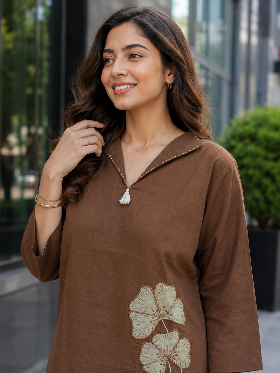 May include: A brown long-sleeved tunic featuring a V-neckline with a small white tassel detail and white contrast stitching along the collar. The lower portion of the garment displays two embroidered light green floral motifs with detailed stitching.
