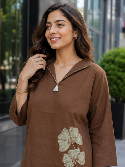 May include: A brown long-sleeved tunic featuring a V-neckline with a small white tassel detail and white contrast stitching along the collar. The lower portion of the garment displays two embroidered light green floral motifs with detailed stitching.