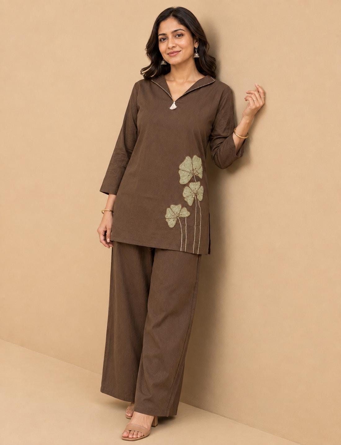 May include: A dark brown tunic and matching wide-leg trousers set. The tunic features a V-neckline with a small white tassel detail and three-quarter length sleeves. A light green embroidered floral design is positioned on the lower left side.