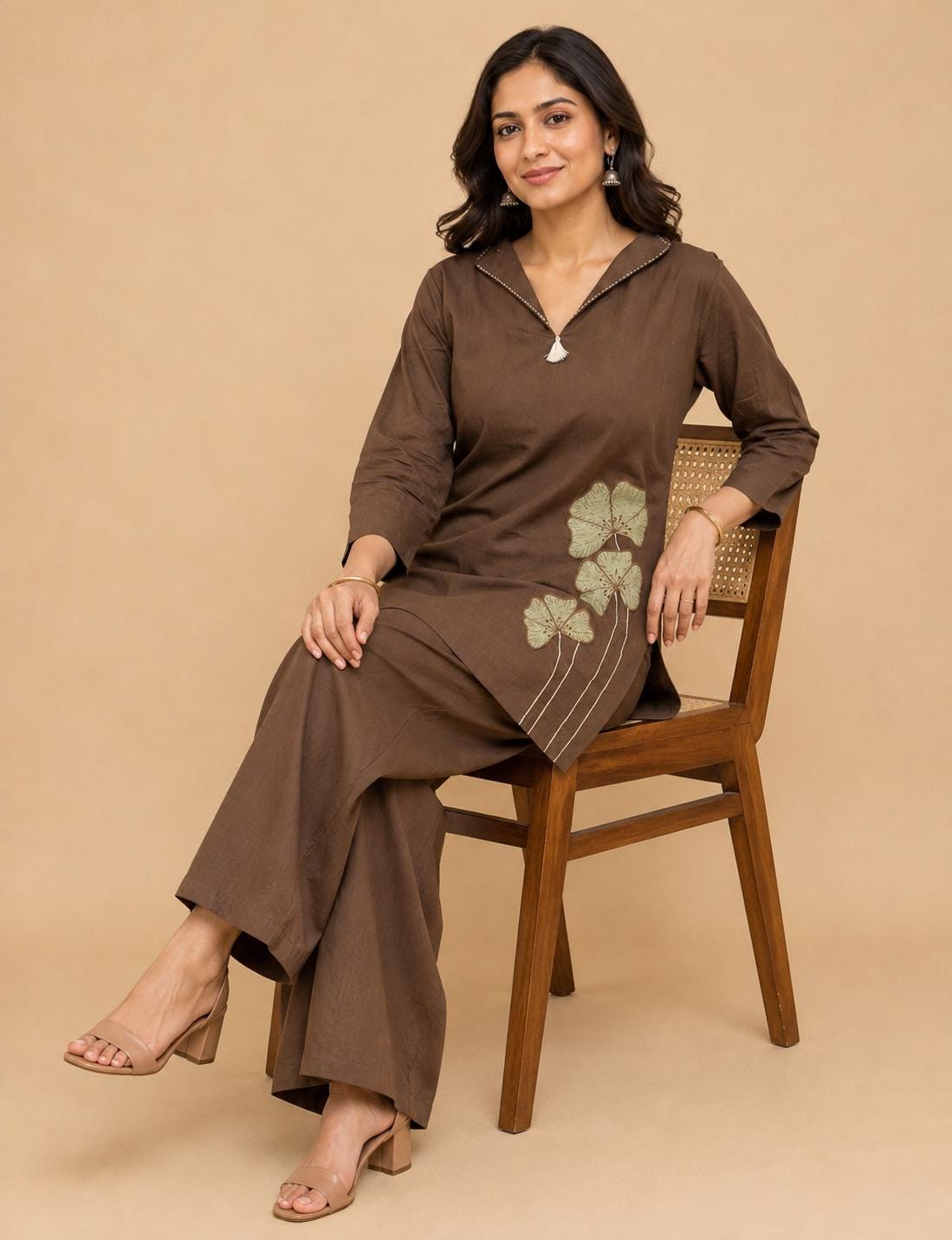 May include: A dark brown long-sleeved tunic with a notched collar and a small white tassel detail, paired with matching wide-leg trousers. The tunic features a light green embroidered floral design on the lower left side with thin stems.