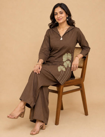 May include: A dark brown long-sleeved tunic with a notched collar and a small white tassel detail, paired with matching wide-leg trousers. The tunic features a light green embroidered floral design on the lower left side with thin stems.