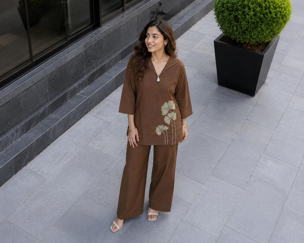 May include: A brown two-piece set consisting of a long-sleeved tunic with a collared V-neckline and wide-leg trousers. The tunic features a light green floral embroidery on the lower left side and a small white tassel detail at the neckline.