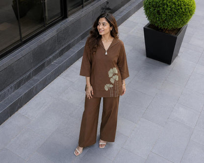 May include: A brown two-piece set consisting of a long-sleeved tunic with a collared V-neckline and wide-leg trousers. The tunic features a light green floral embroidery on the lower left side and a small white tassel detail at the neckline.