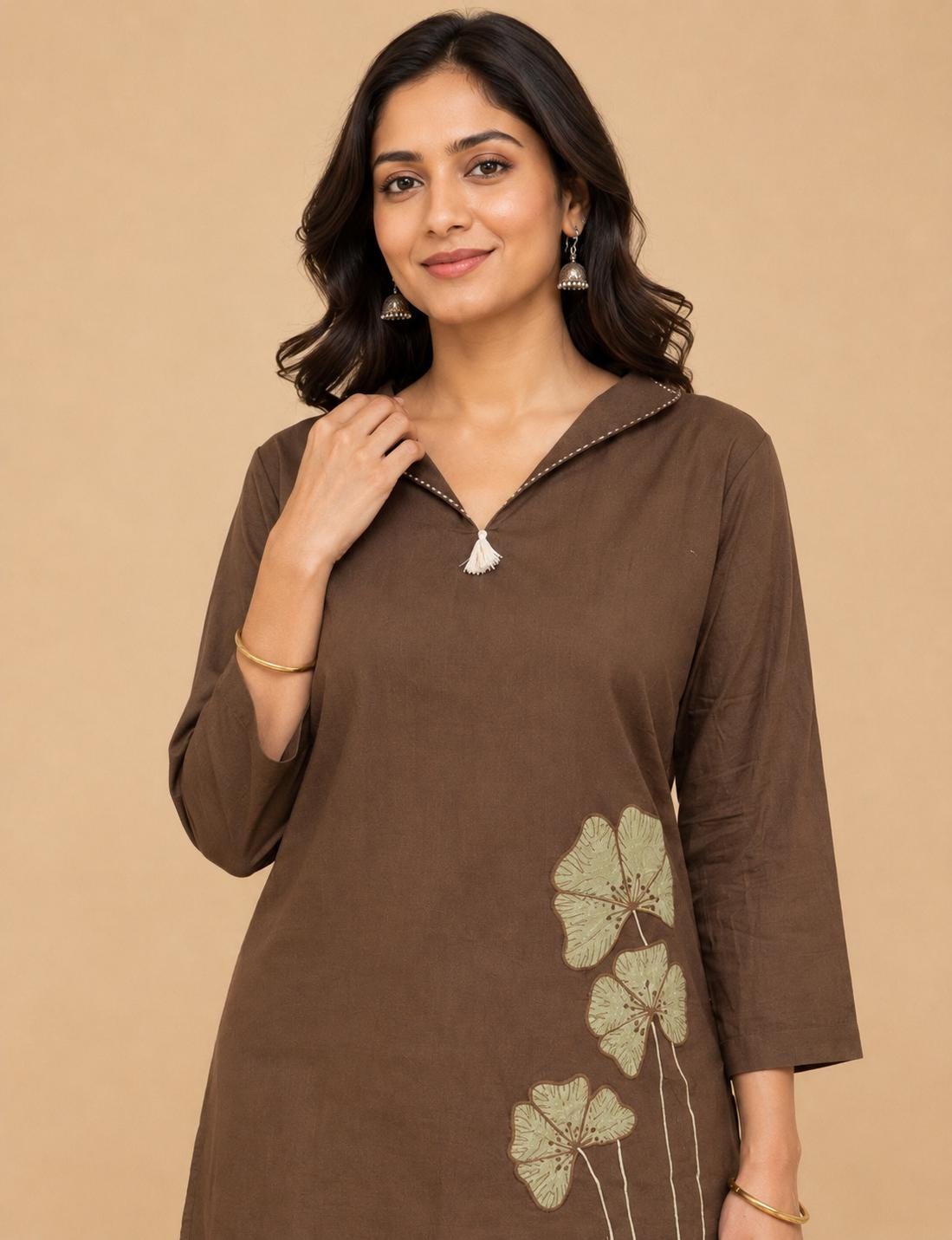 May include: A dark brown long-sleeved tunic featuring a V-neckline with a small white tassel detail and light green embroidered floral motifs on the lower side. The garment has a simple collar and straight sleeves, providing a clean look.